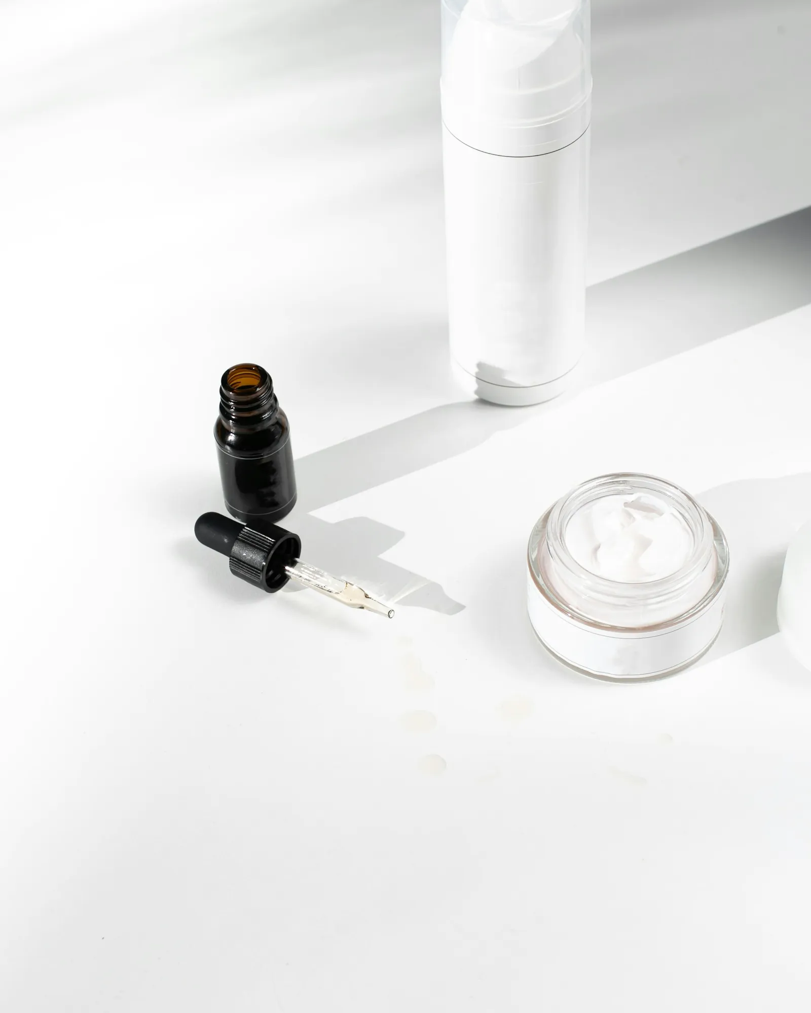 Amber dropper bottle, white airless bottle and cream jar on a white surface